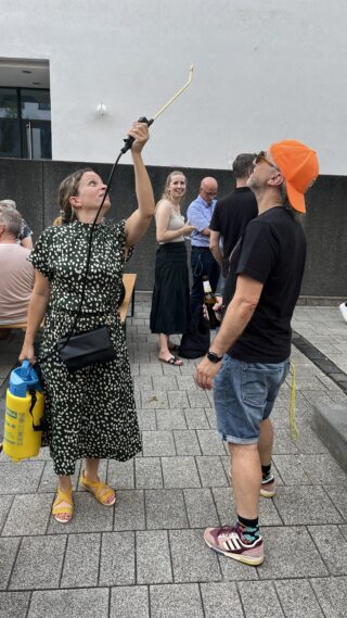 Eine Person steht unter einem tragbaren Wassersprüher, welcher von einer Frau gehalten wird.